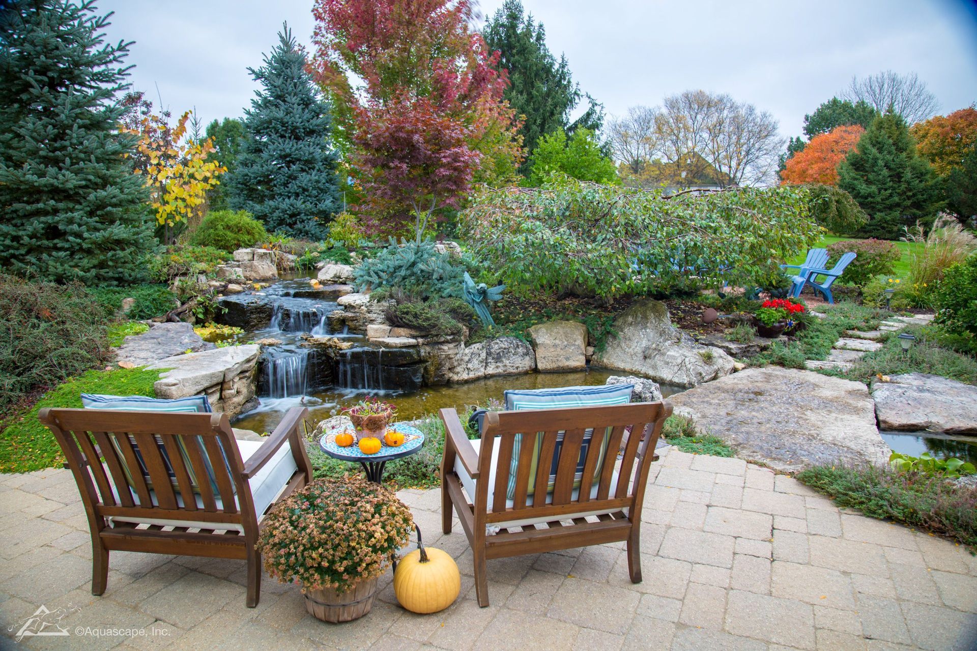 Two wooden chairs face a small waterfall and pond in an autumn garden, pumpkins and foliage present.