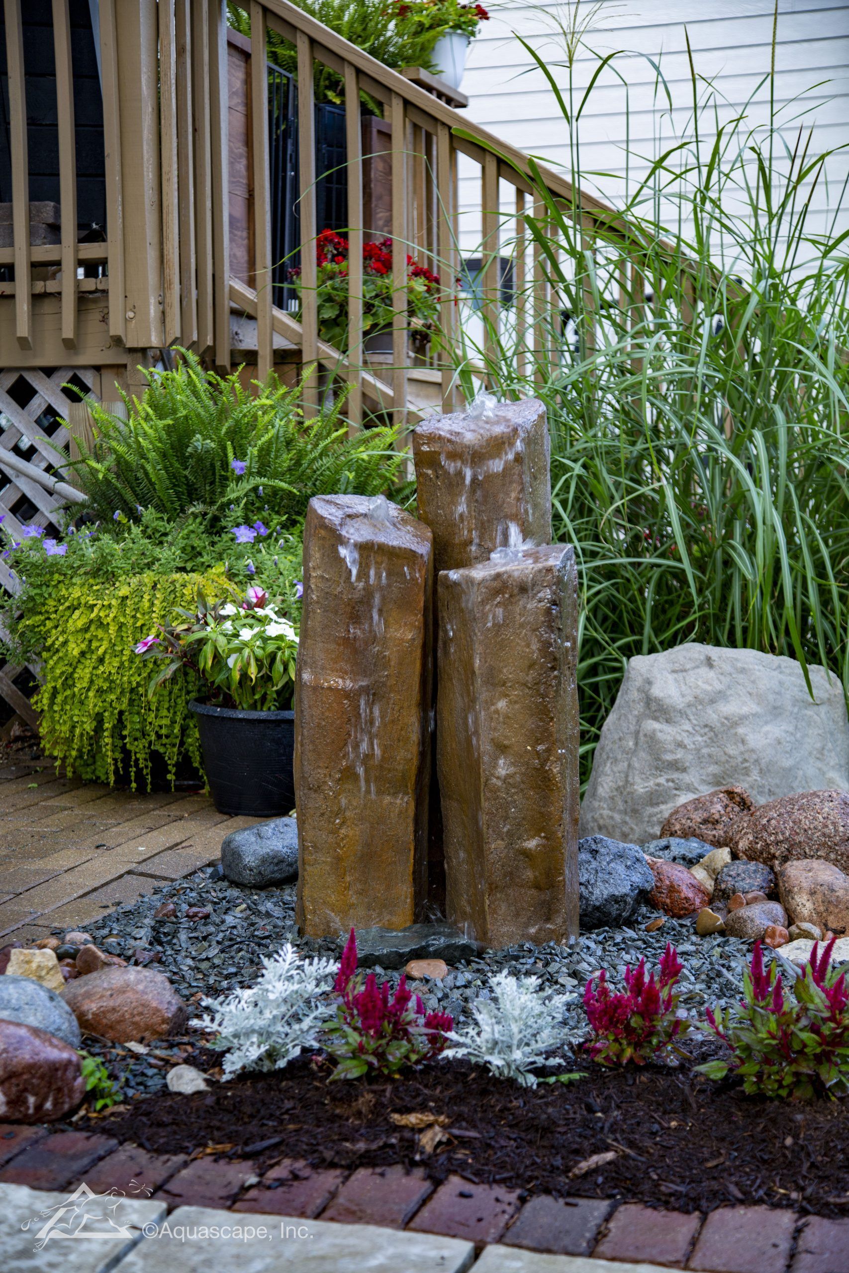 Water fountain with three rectangular stone pillars; surrounded by plants, rocks, and a wooden deck.