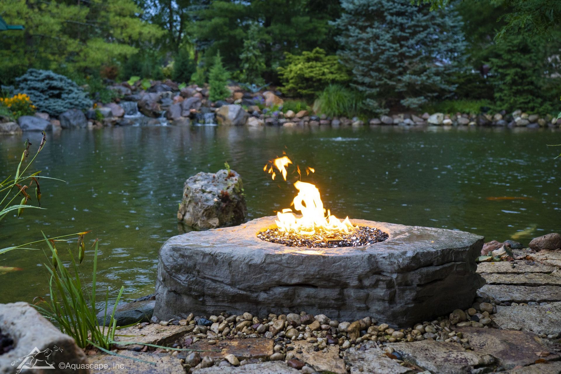 Stone fire pit with flames, beside a pond surrounded by trees and rocks.