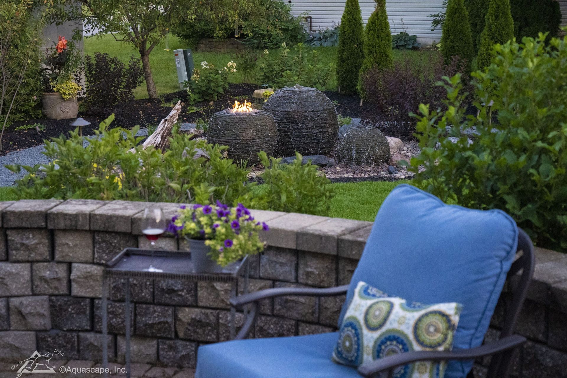 Patio with stone wall and blue chair, overlooking a garden with water feature and small fire.