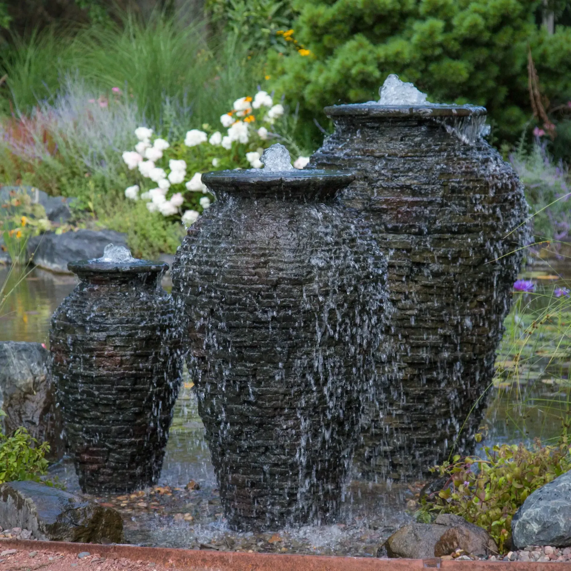 Three dark, textured urn fountains with water cascading into a pond, surrounded by plants.
