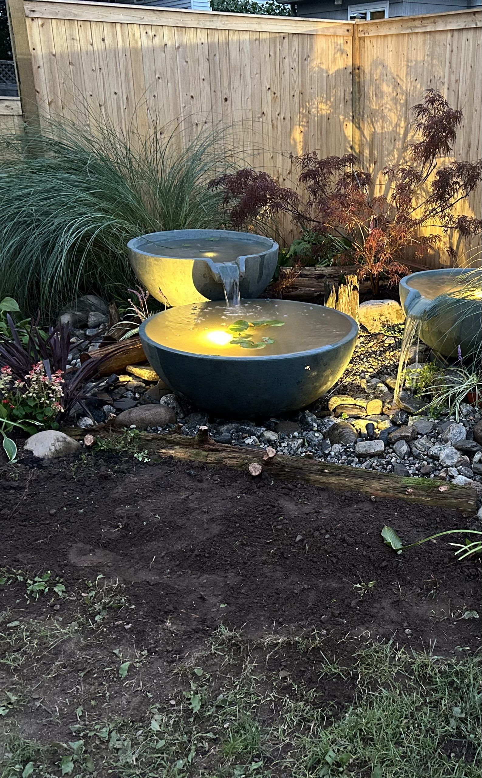 Water fountain in a garden. Three stone bowls, water flows, lit from below. Behind, a fence and plants.