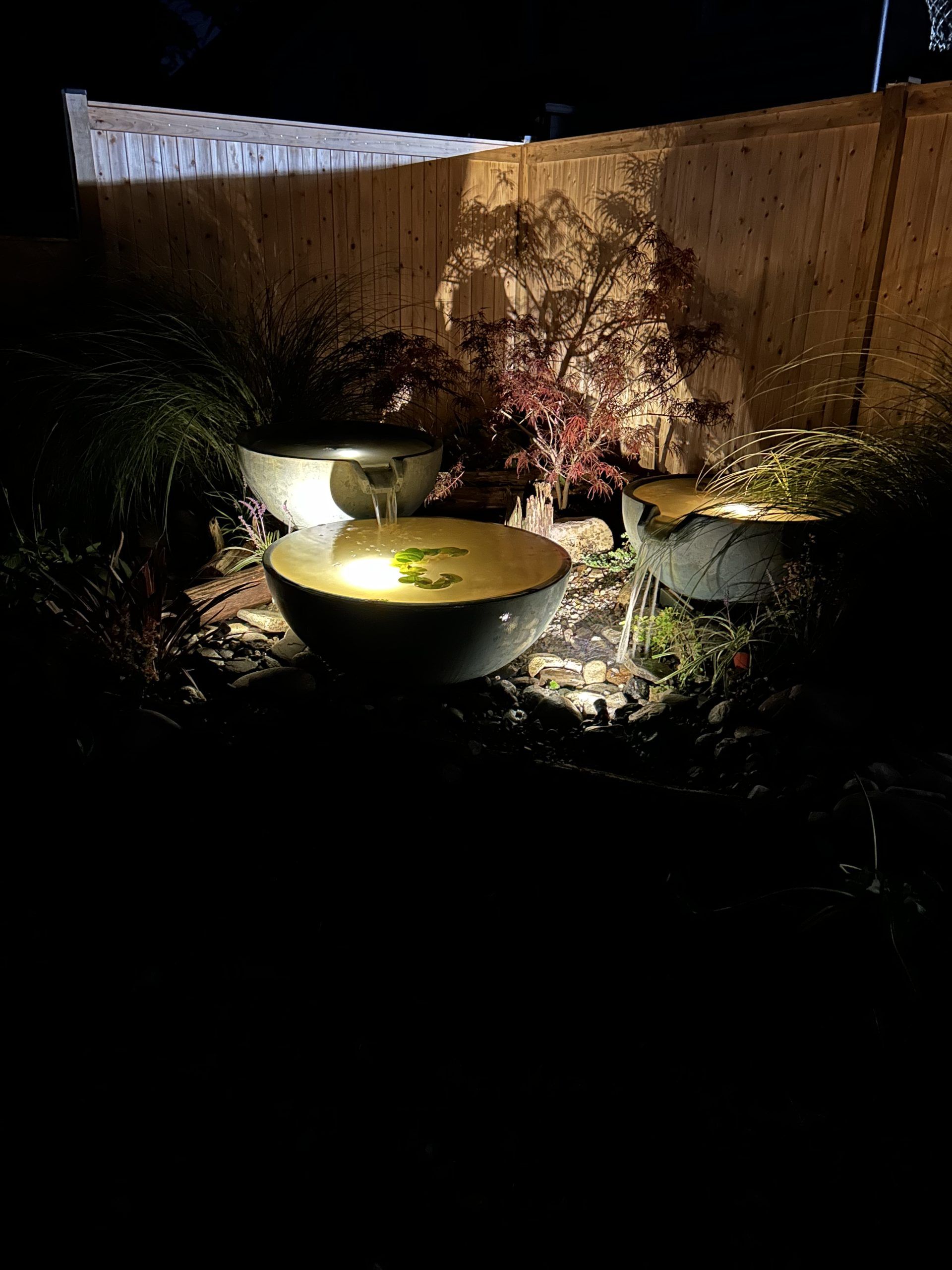 Nighttime view of a water feature with three stone bowls illuminated by a spotlight, plants, and a bamboo fence in the background.