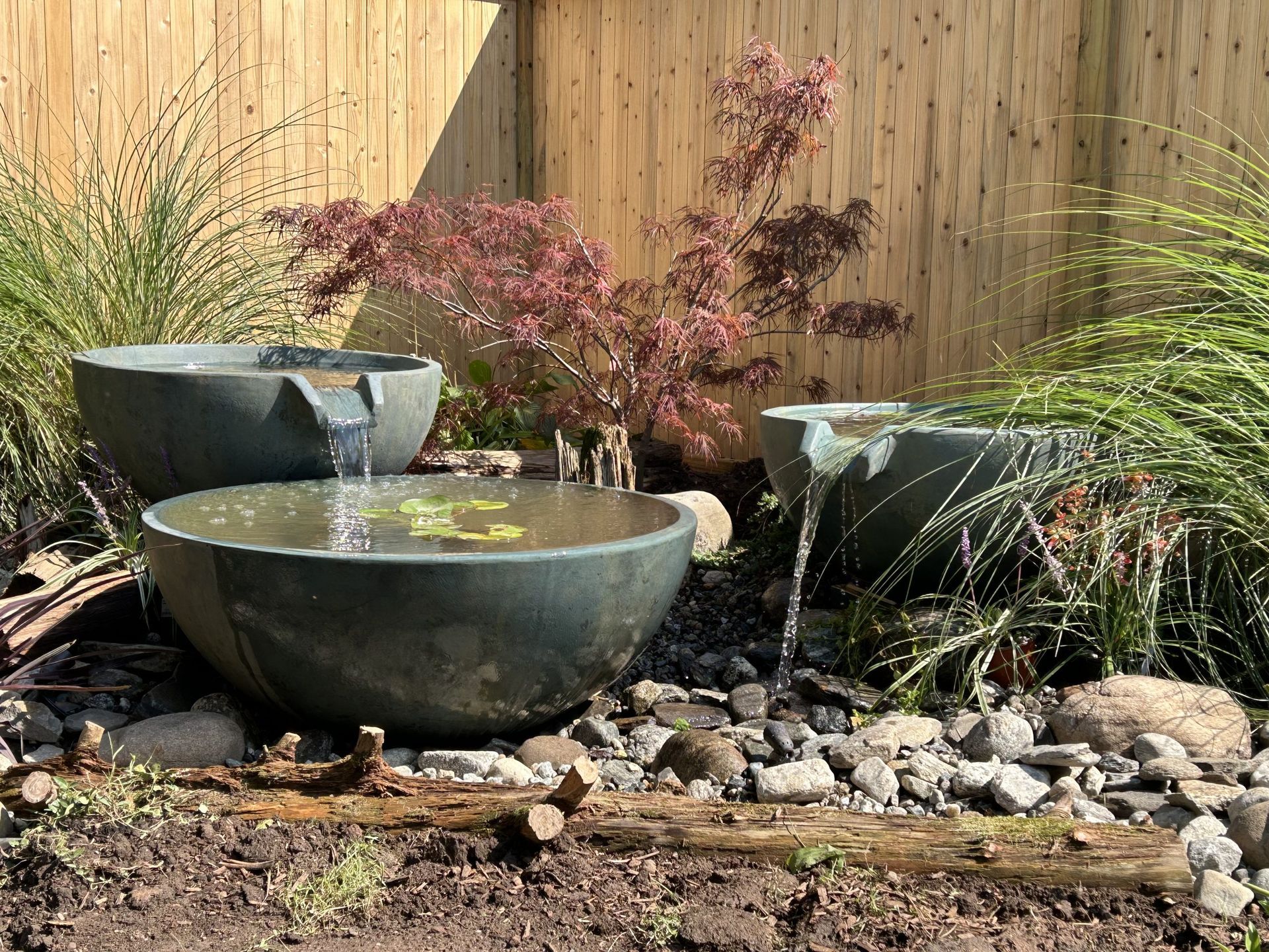 Stone water fountain with three basins, surrounded by rocks, plants, and a wooden fence background.