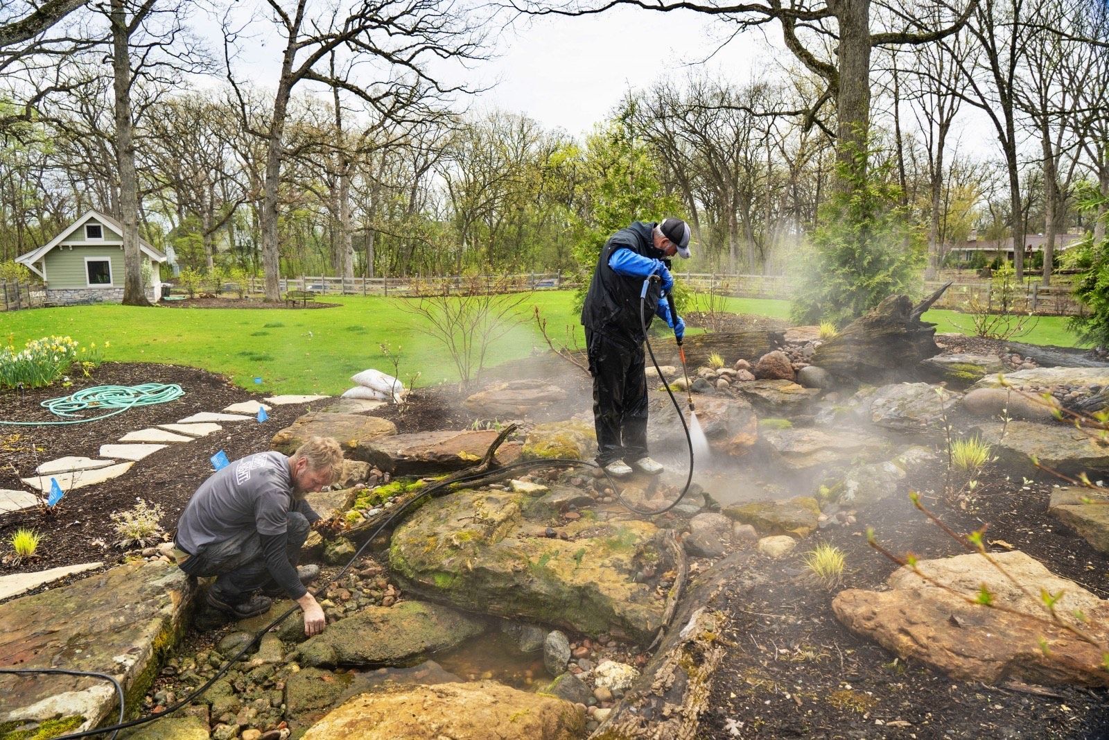 Two people cleaning a garden pond with a hose, mist rising. Lush green lawn, trees, and a house in the background.