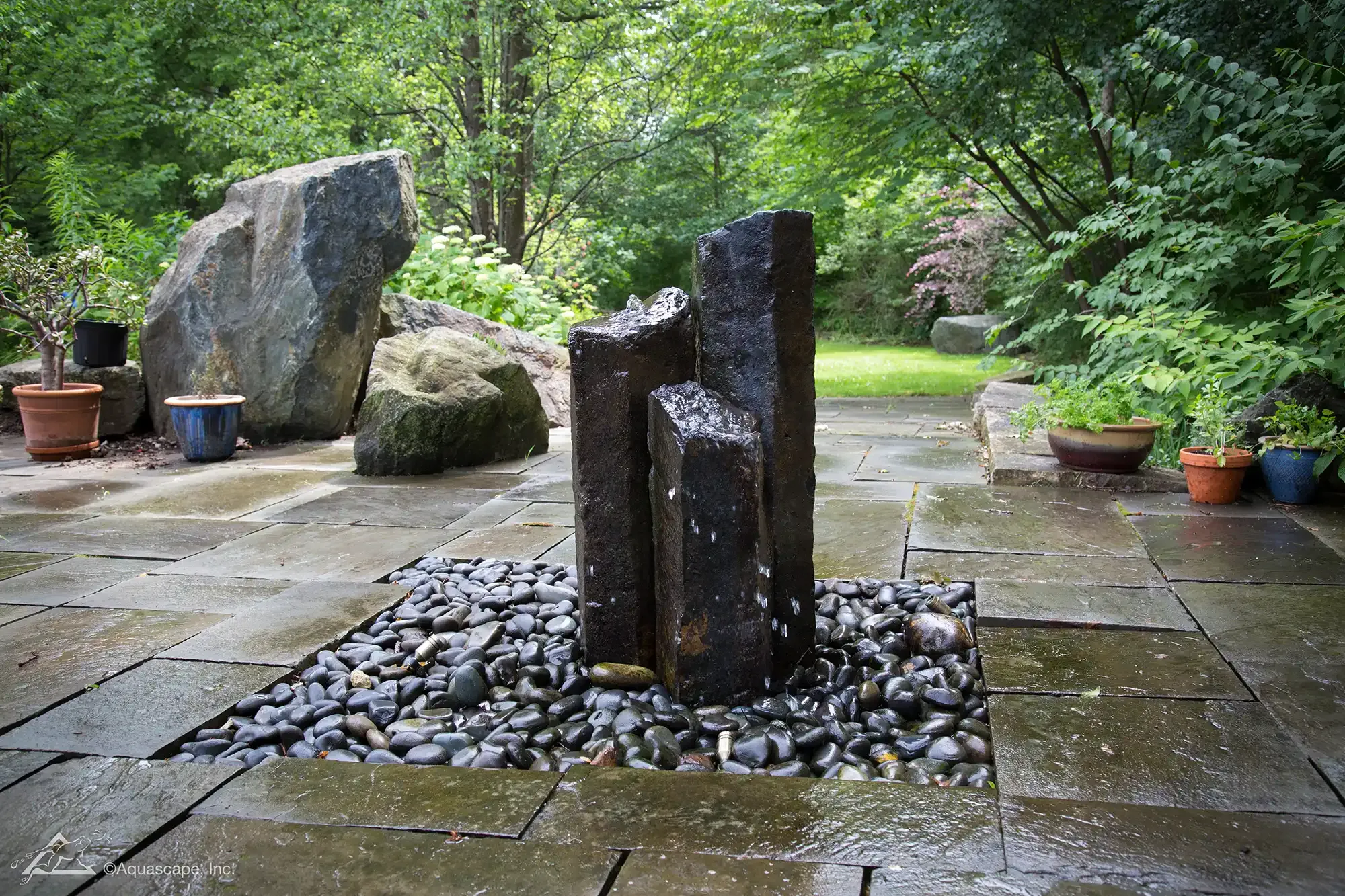 Stone fountain on a dark stone patio surrounded by greenery and large boulders.