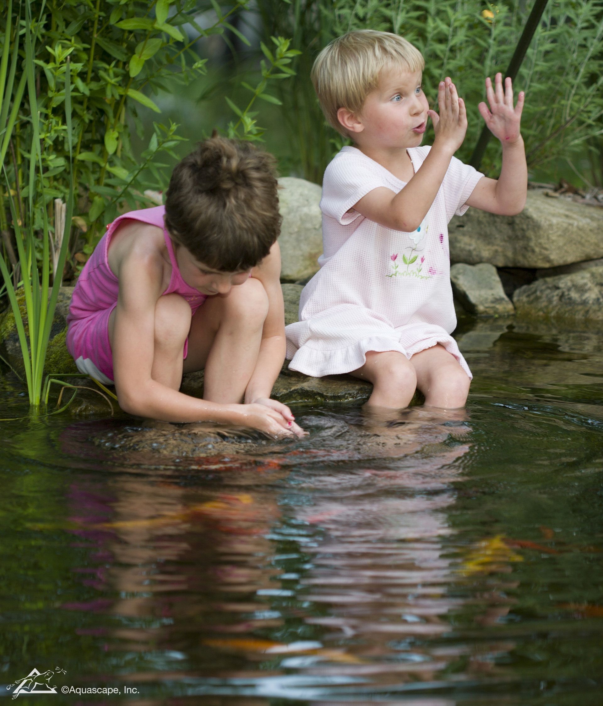 Two children sit by a pond. One looks at water, the other gestures with hands.