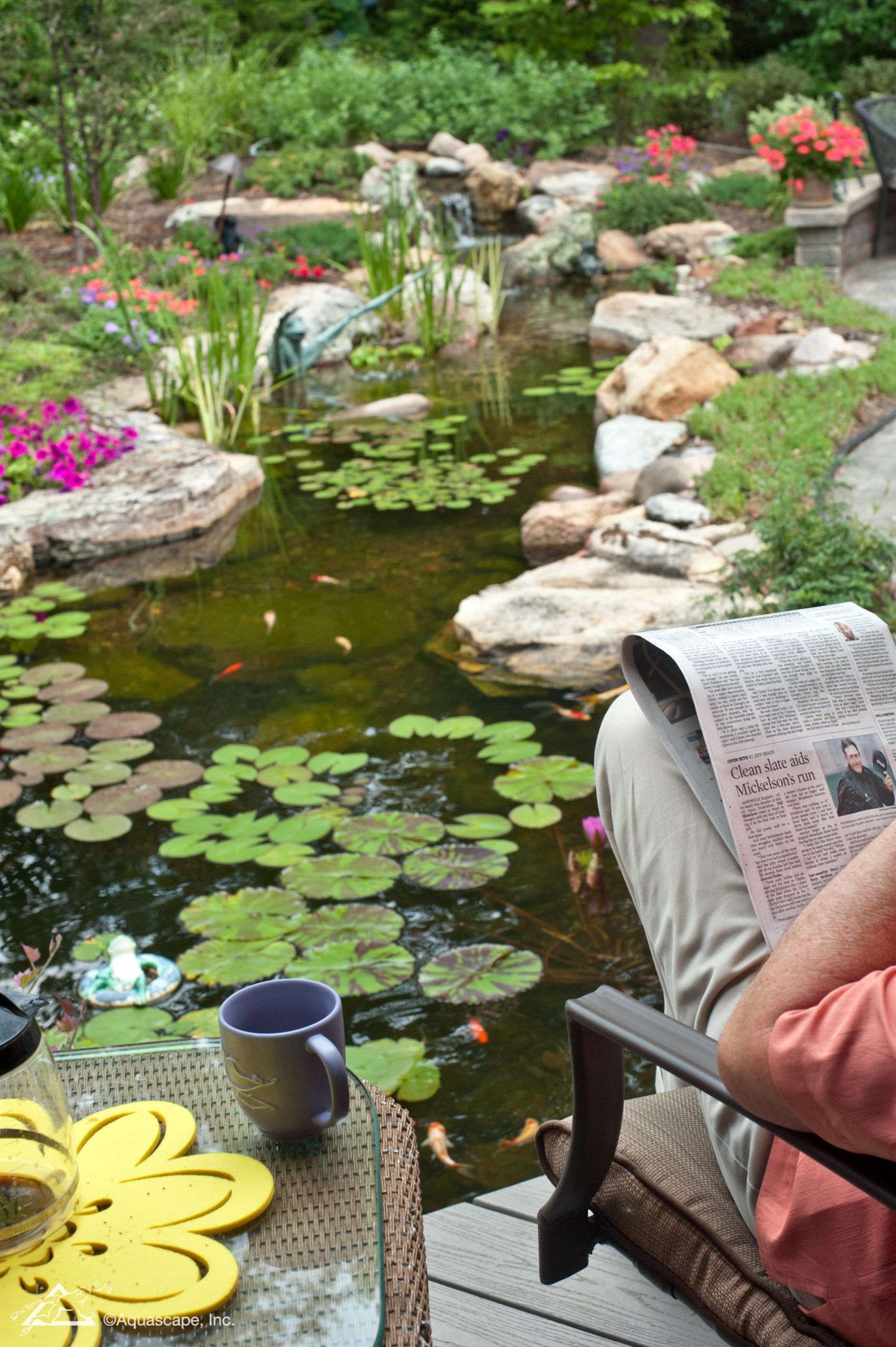 Person relaxing by a pond reading newspaper, cup on table. Lily pads, flowers and waterfall in the background.