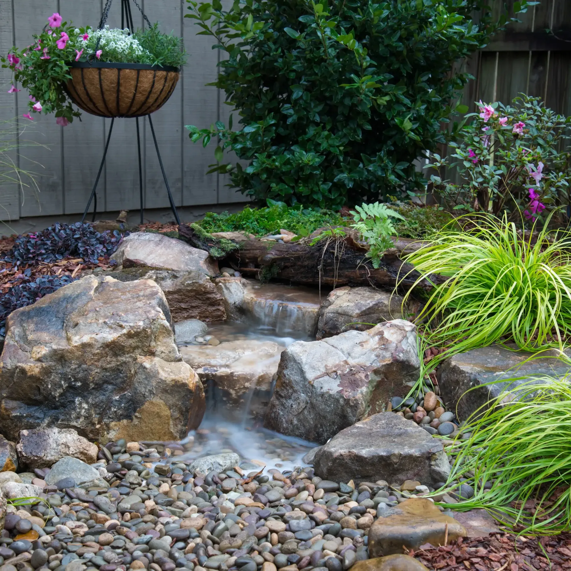 Small backyard waterfall with rocks, plants, and hanging basket of flowers.