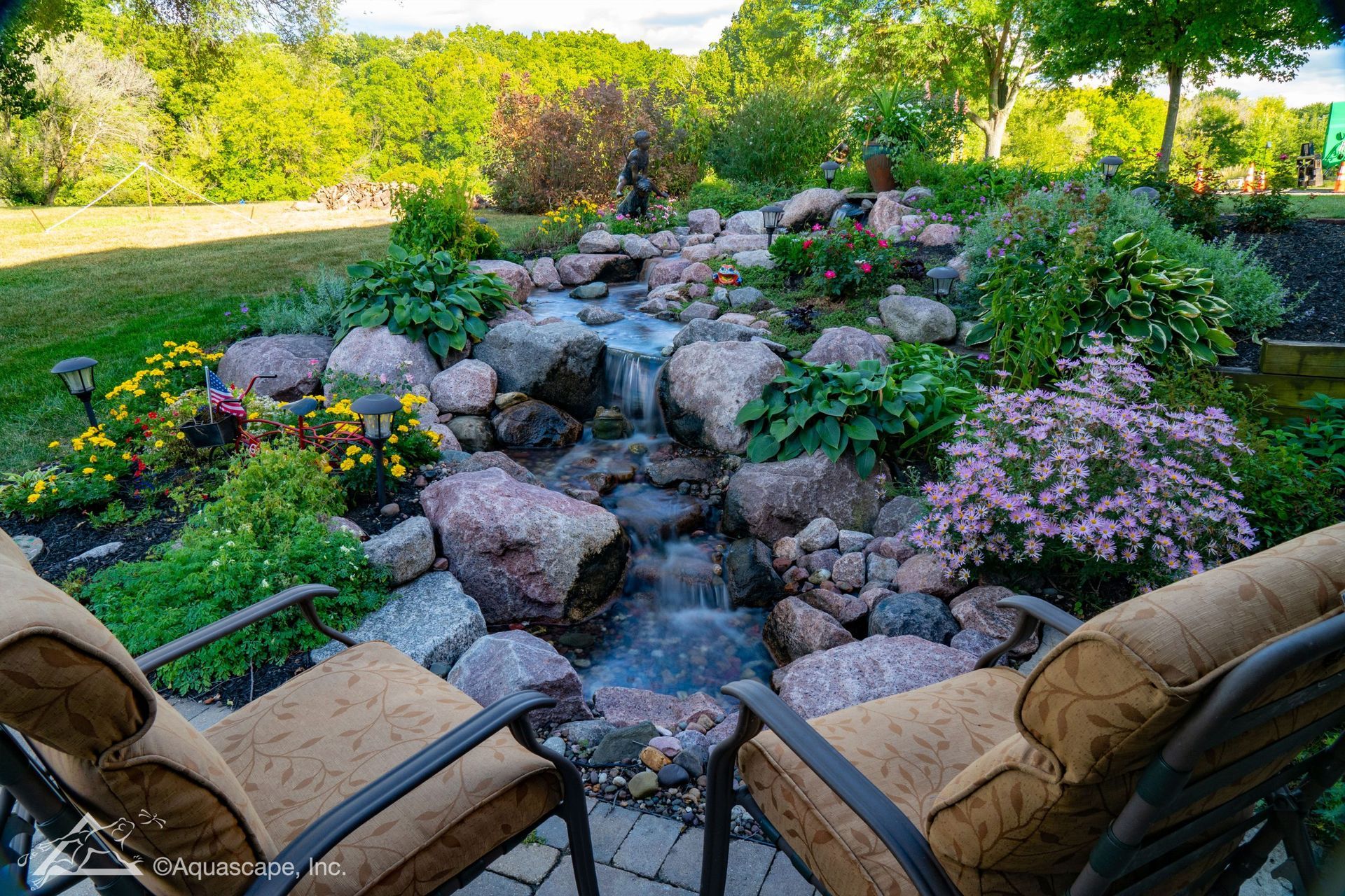 Two patio chairs overlook a lush garden with a flowing waterfall surrounded by rocks and colorful flowers.