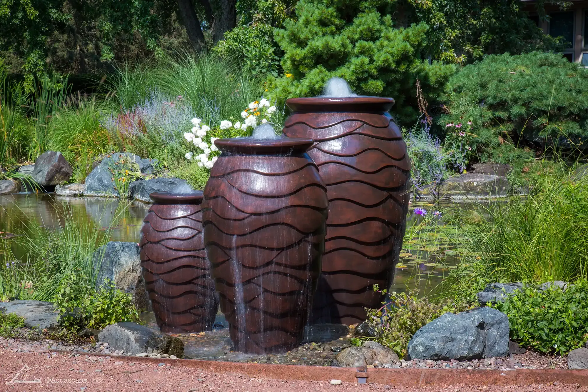 Three brown urn water fountain feature in a garden, water flowing, with pond and greenery.