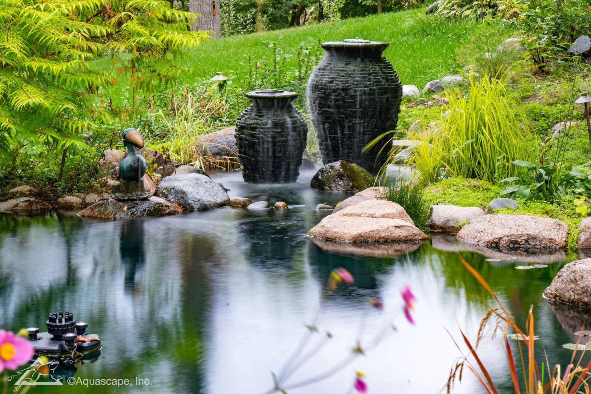 Pond with water feature: three dark urns spout water into a reflective pool surrounded by rocks and lush greenery.