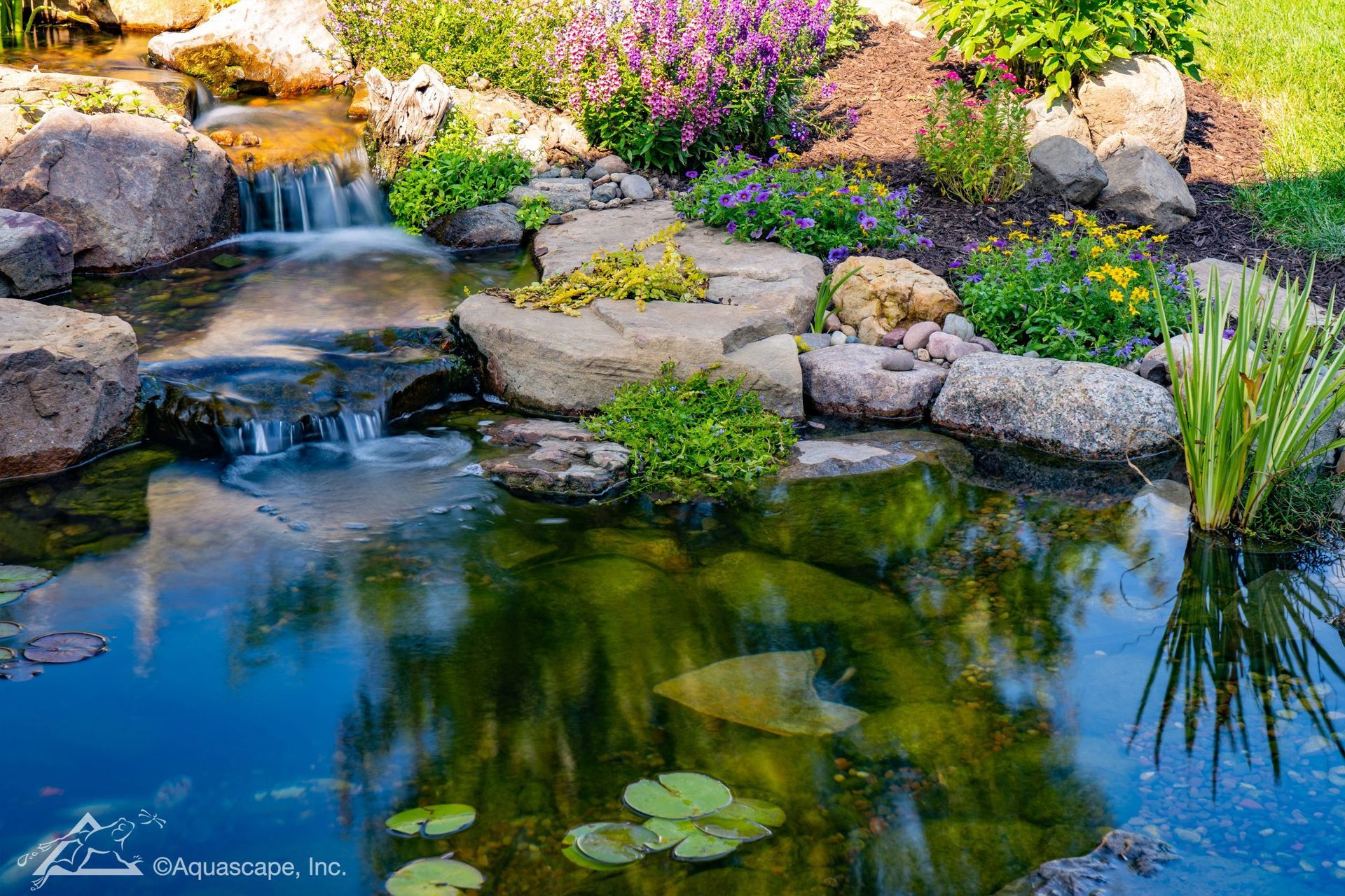 Waterfall flows into a pond surrounded by rocks and colorful flowers.