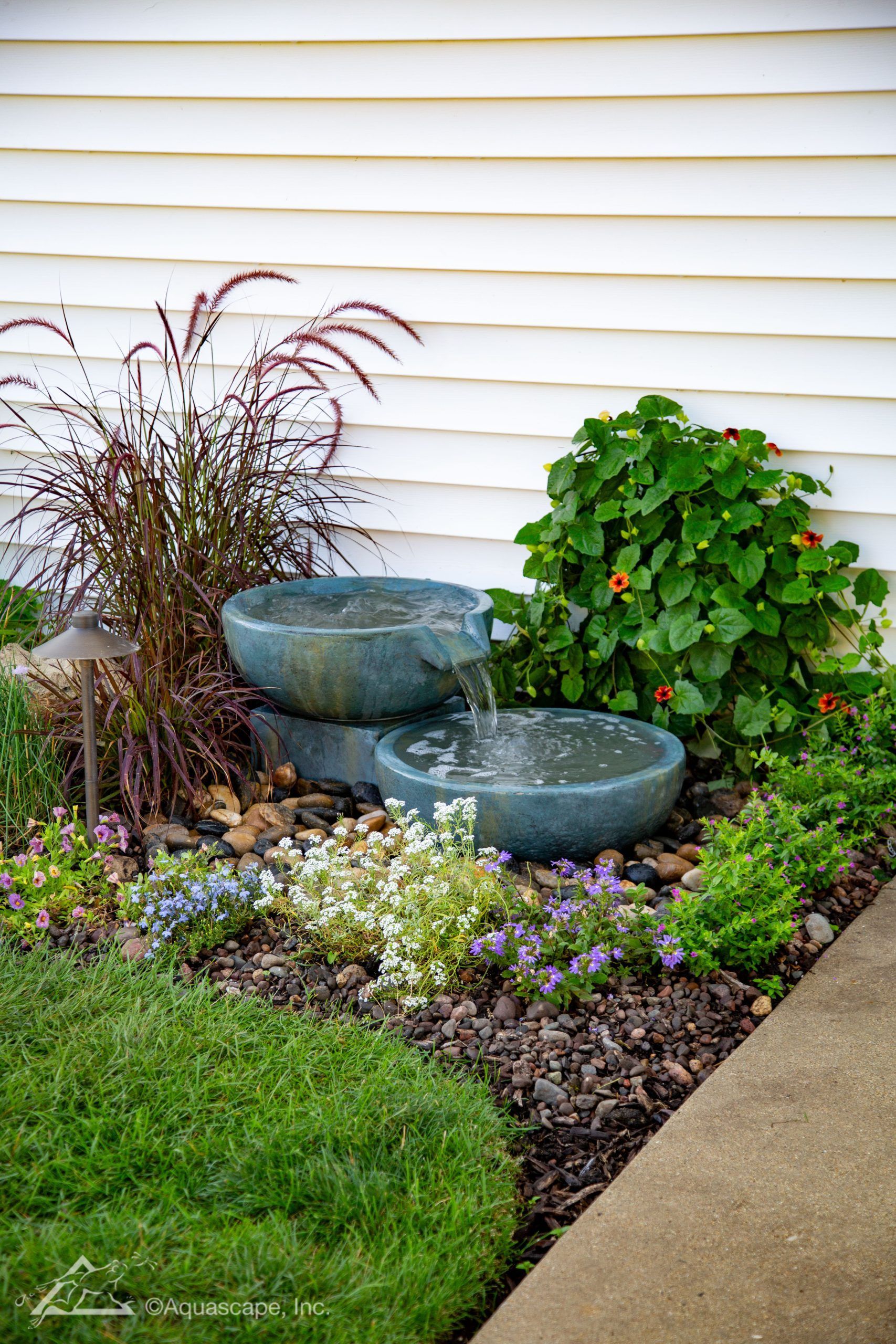 Water fountain in a garden bed with lush greenery and colorful flowers against a white building.
