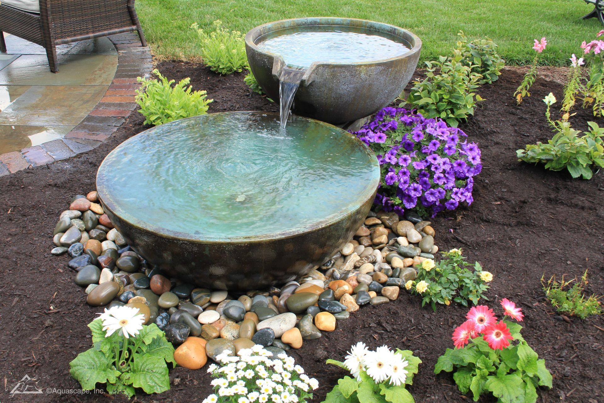 Water fountain with two bowl-shaped basins, overflowing into a bed of river rocks and surrounded by flowers.