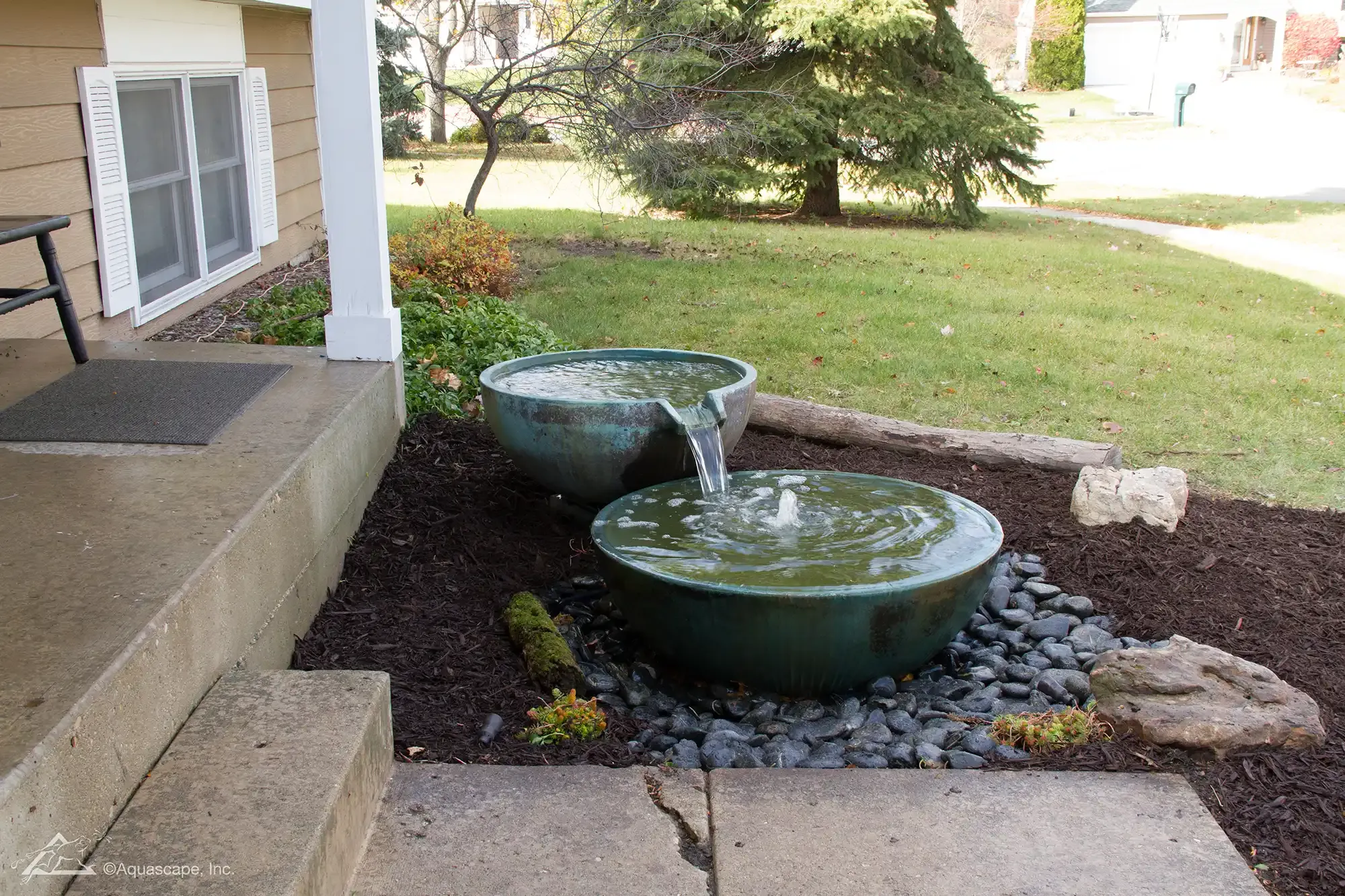 Water fountain feature on a porch, consisting of two green ceramic bowls, water flowing over black rocks and mulch.