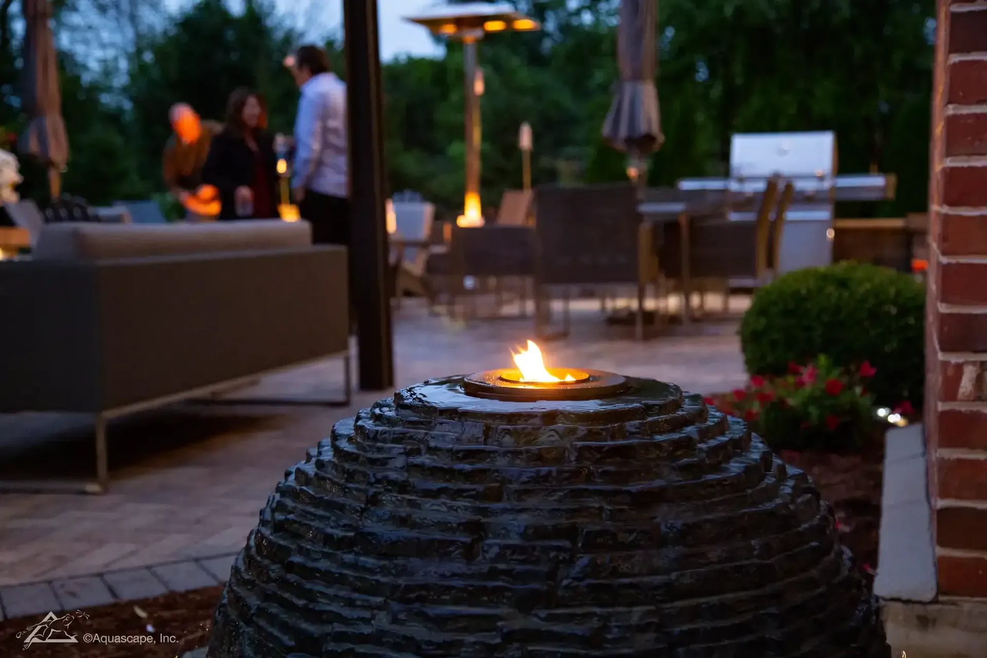 Stone fire pit with flames, outdoor patio with people, grill, furniture, and heaters.
