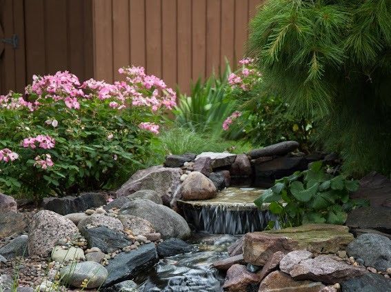 Small waterfall cascading over rocks in a garden, with pink flowers and green plants. Brown fence in background.