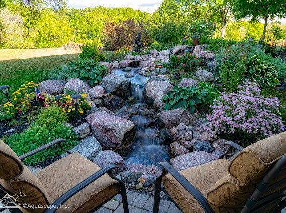 Two chairs face a rocky waterfall in a lush garden with colorful flowers and trees.