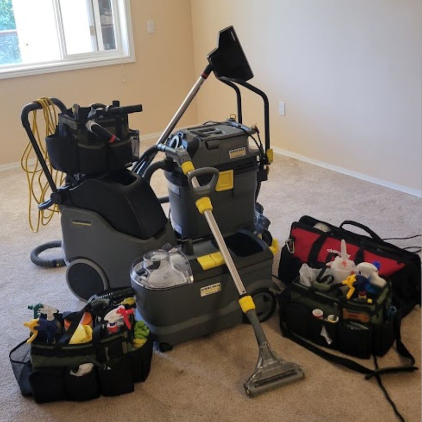 Carpet cleaning equipment: large machines, accessories, and bags on beige carpet in a room with a window.