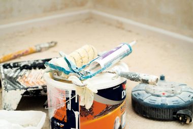 Paint supplies on floor: paint can, rollers, tray, and extension cord near a wall.