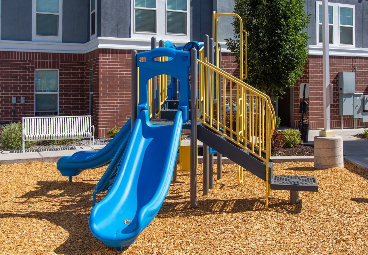 Blue playground with slides and yellow railings in a courtyard beside an apartment building.