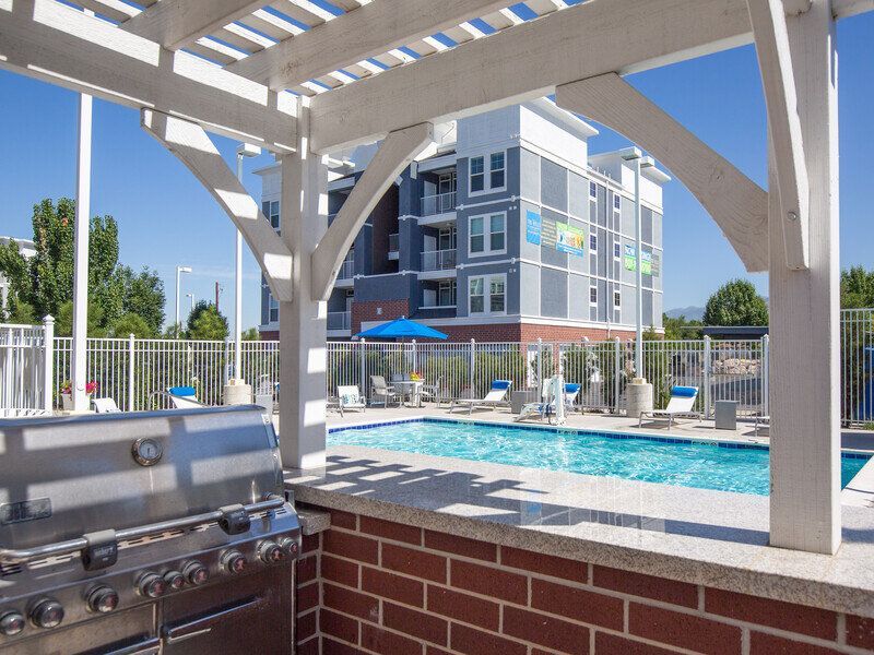 Outdoor pool area with a grill under a pergola and lounge chairs, with an apartment building in the background.