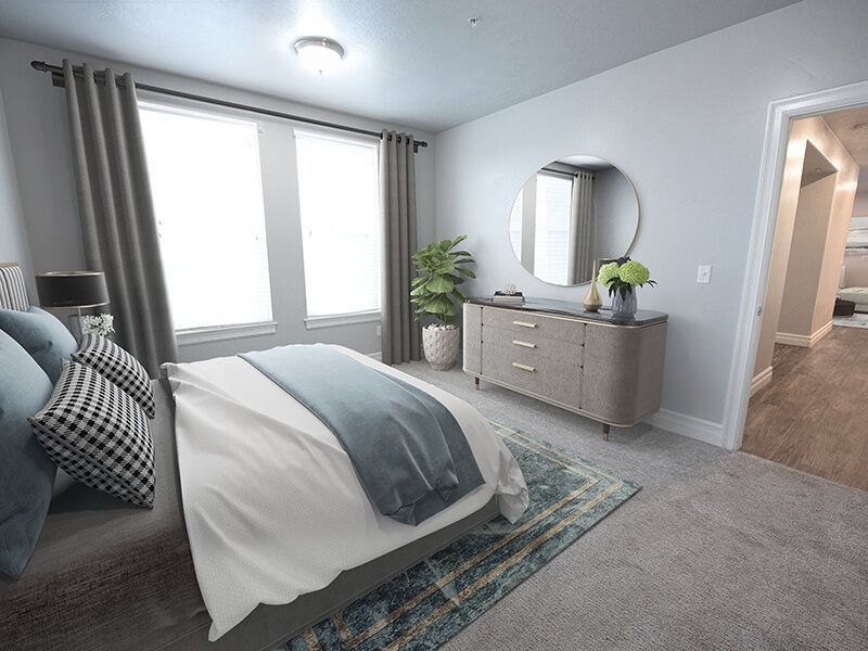Bedroom in a modern apartment with a king bed, grey dresser, round mirror, potted plant, and two light-filled windows.
