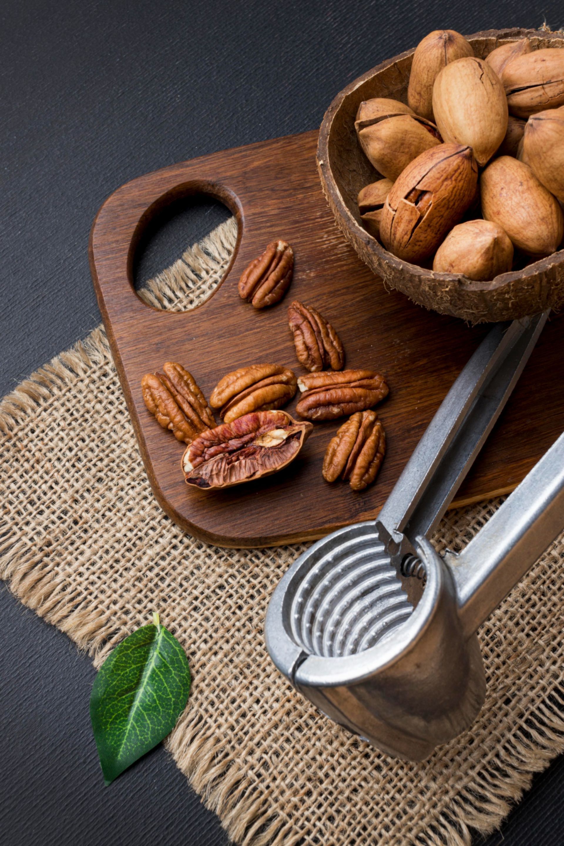 Pecan nuts on a wooden board, with a coconut shell, nutcracker, and burlap, against a dark background.