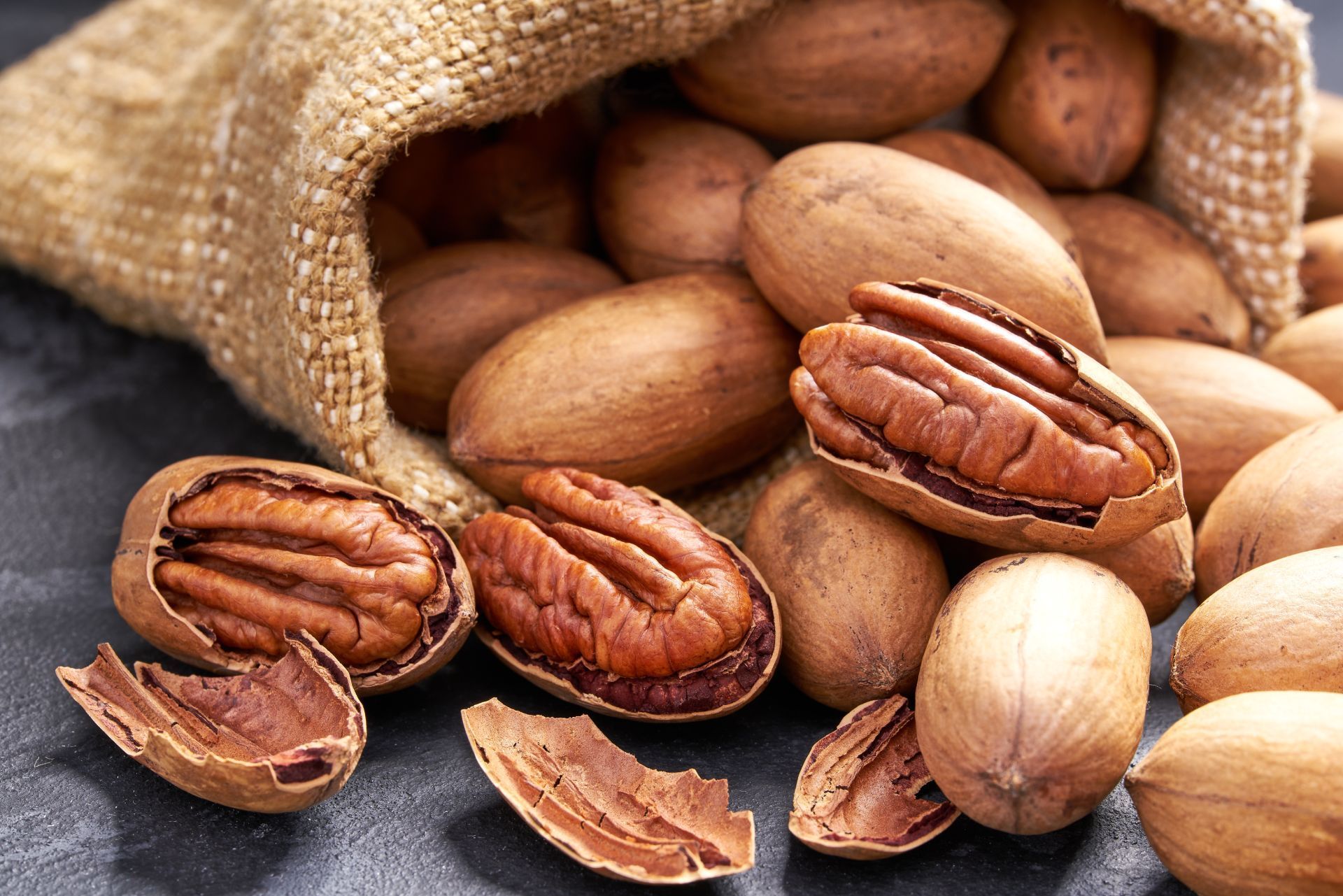 Pecans spilling from a burlap sack onto a dark surface. Some are shelled, showing brown nutmeat.