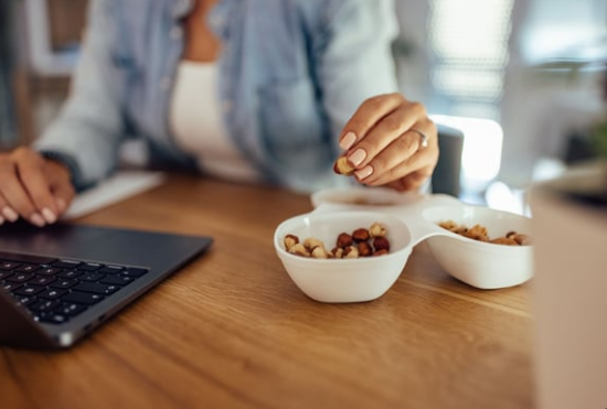 Person eating nuts from a divided dish while using a laptop on a wooden surface.