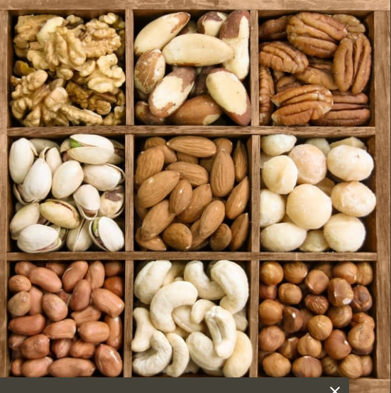 Assorted nuts arranged in a wooden box, including walnuts, Brazil nuts, pecans, pistachios, almonds, macadamia nuts, peanuts, cashews, and hazelnuts.