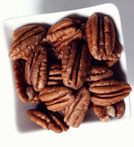 Pecan halves in a white square bowl, close-up shot. Brown nuts, bright lighting.