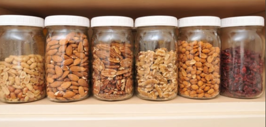 Six glass jars filled with various nuts and dried fruit on a wooden shelf. Each jar has a white lid.