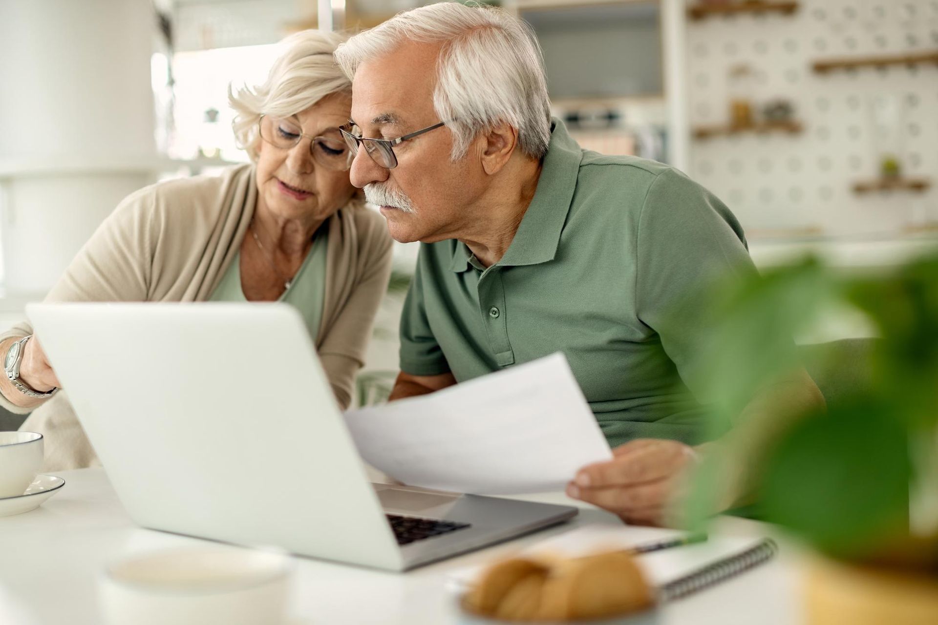 Elderly couple reviewing documents on a laptop at a kitchen table with a coffee cup and plate nearby