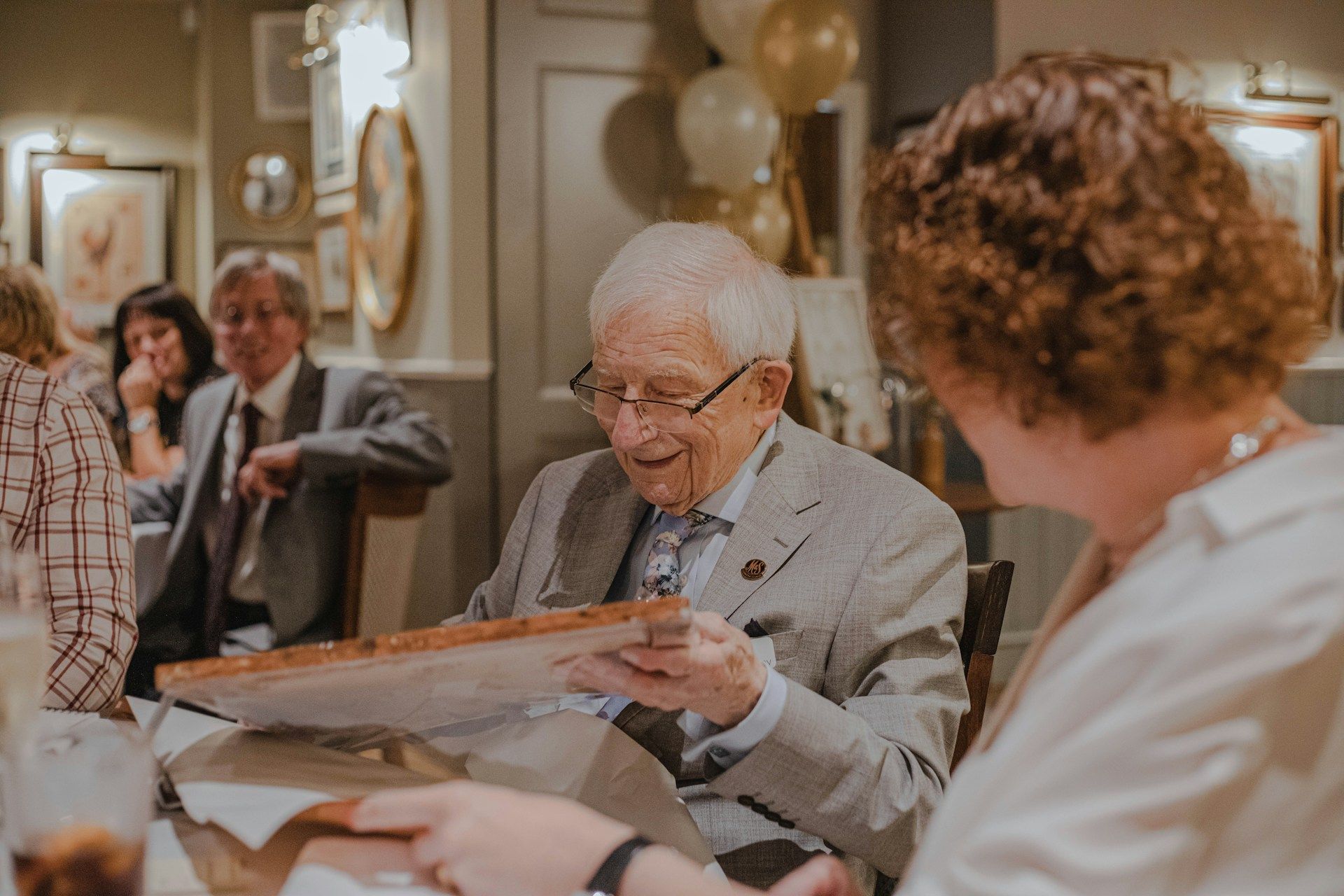 People seated at a festive table, smiling and chatting while an elderly man reads a card.