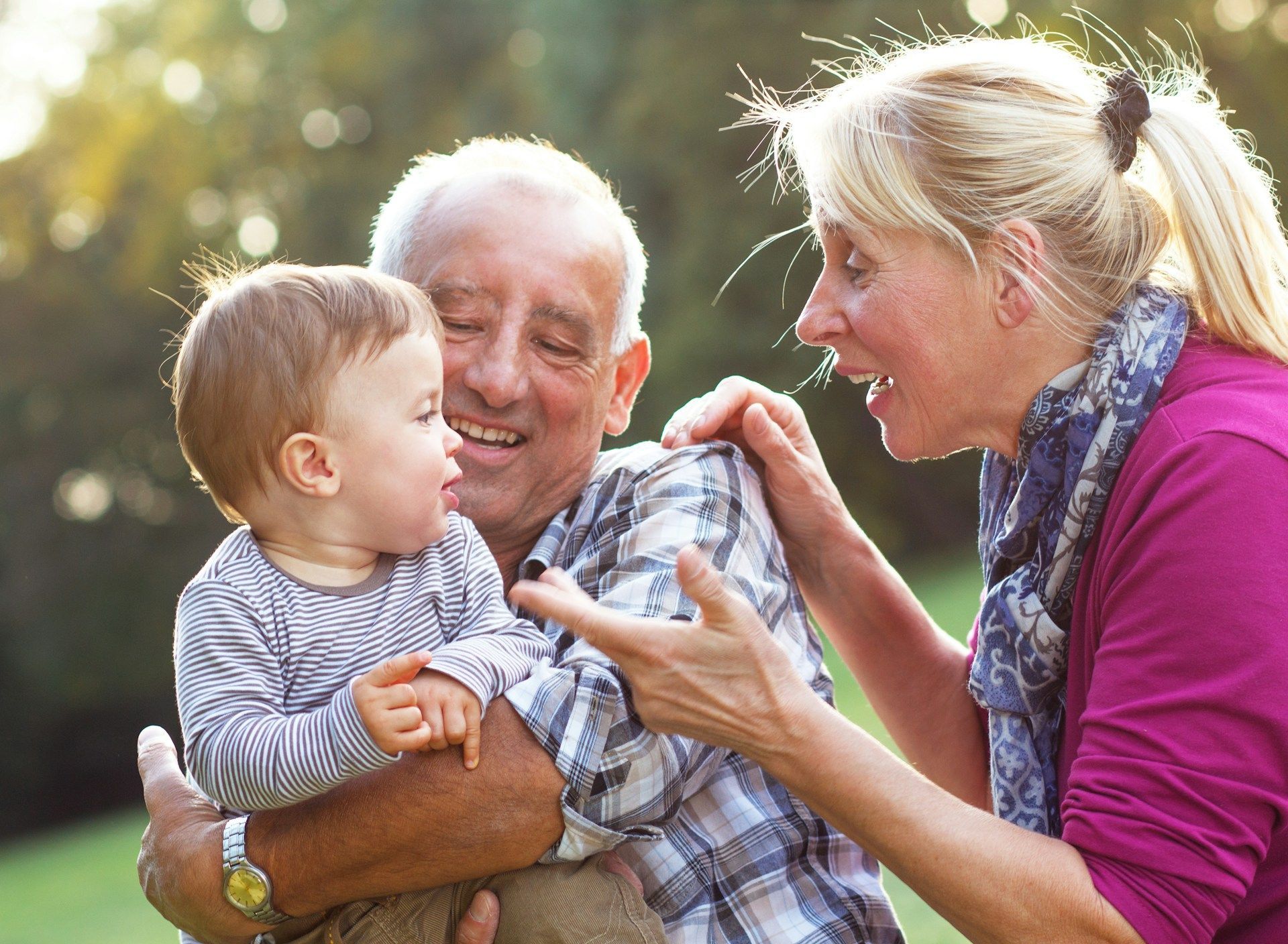 Older man holding a toddler while a blonde woman smiles and reaches toward the child outdoors