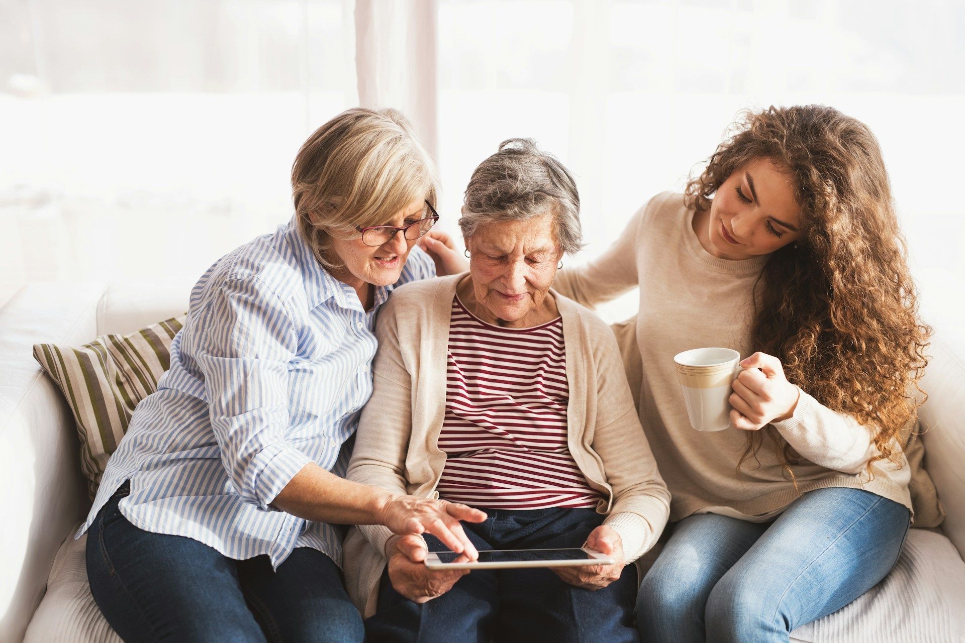 Three people sit on a couch, smiling and looking at a tablet, with one holding a mug.