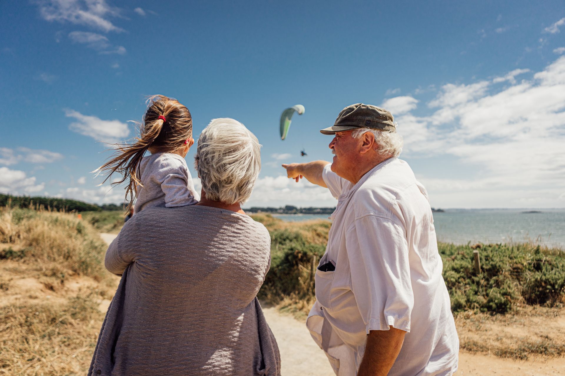 Three people at a windy beach overlook, one holding up a gull-shaped kite against a blue sky.