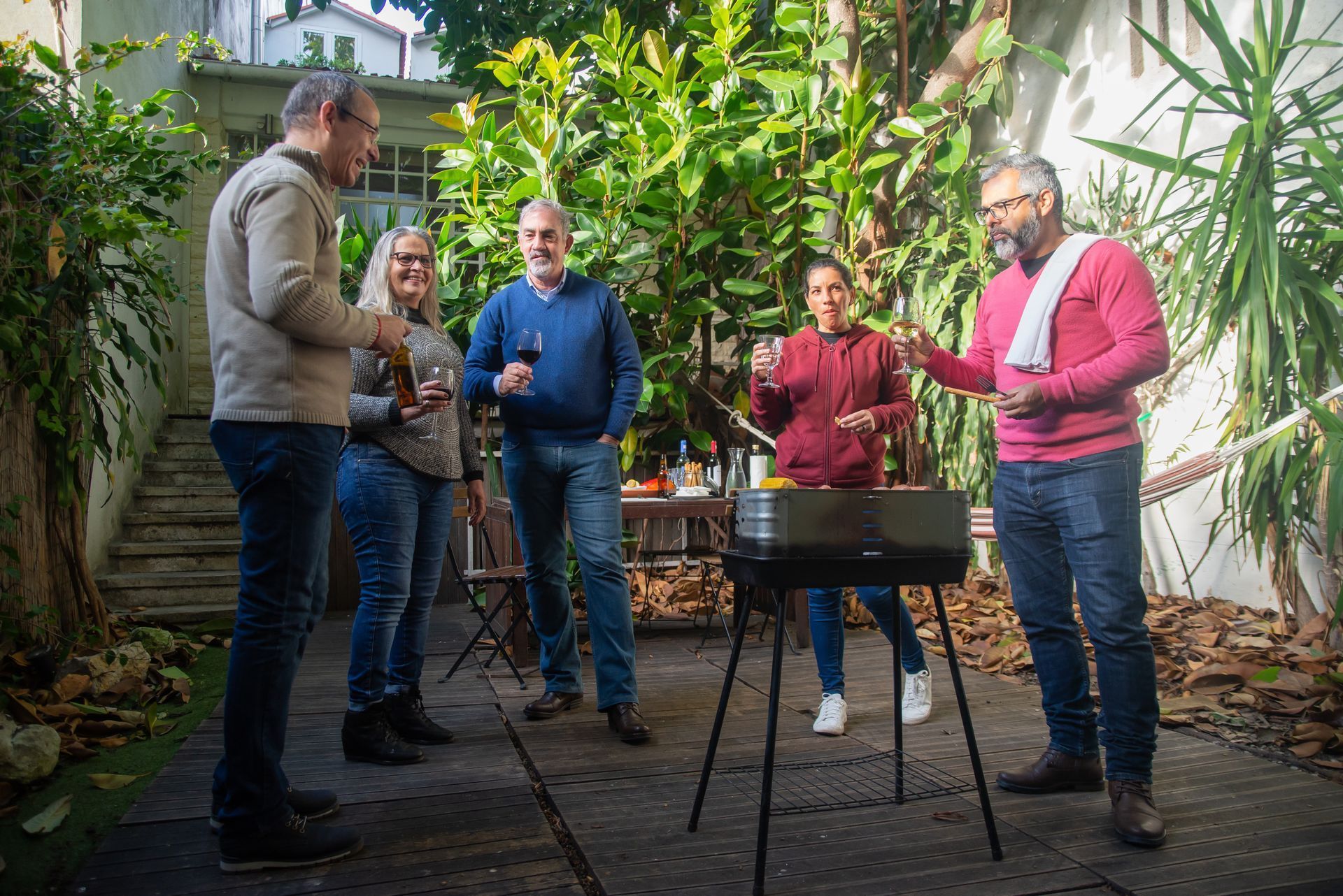 Five people stand around a small fire pit in a lush garden, chatting and holding drinks.
