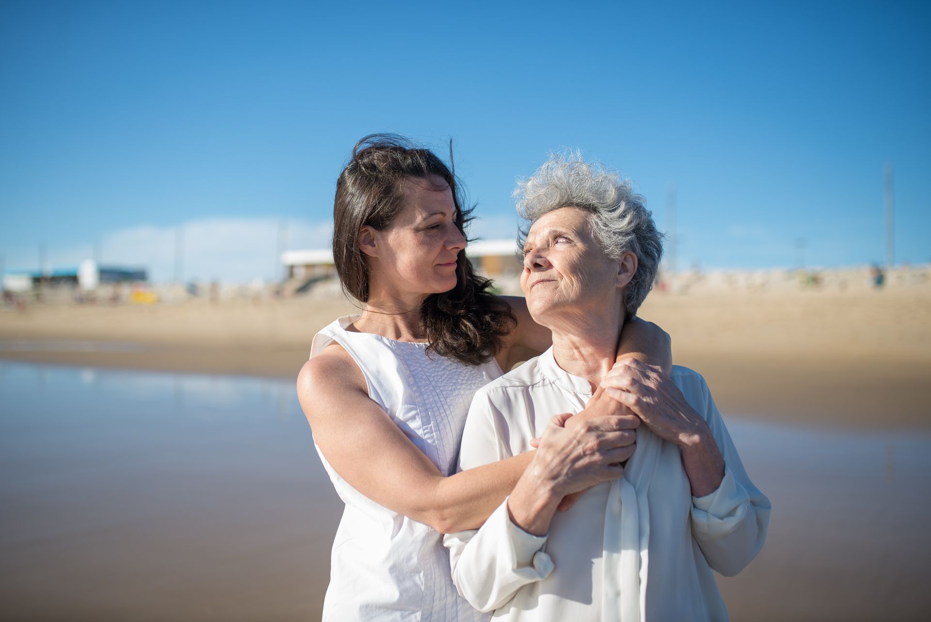 Two people in white clothing stand on a sunny beach, holding hands and smiling at each other.