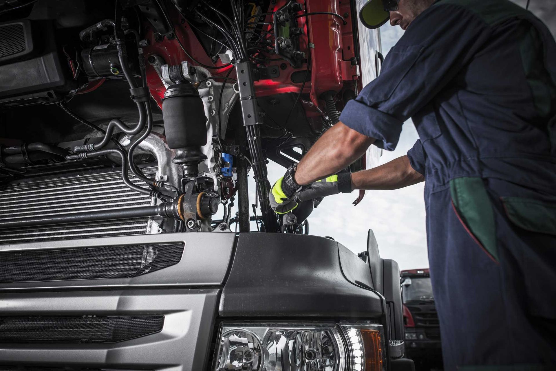 Technician working under an open truck cab, repairing engine components with tools.