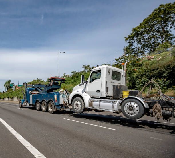 Tow truck towing white semi-truck on highway with trees in background.
