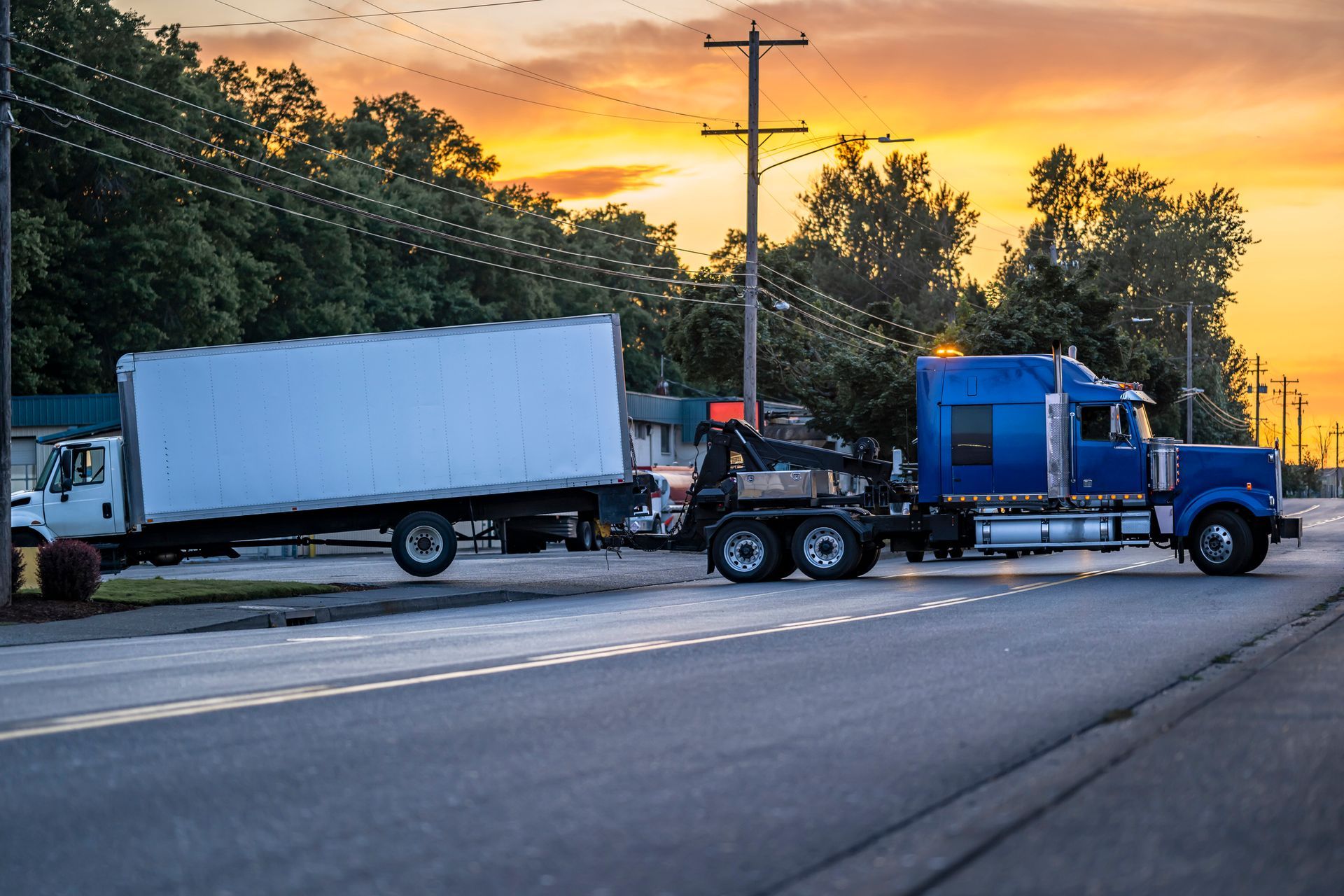Tow truck hauling white box truck at sunset on suburban road.