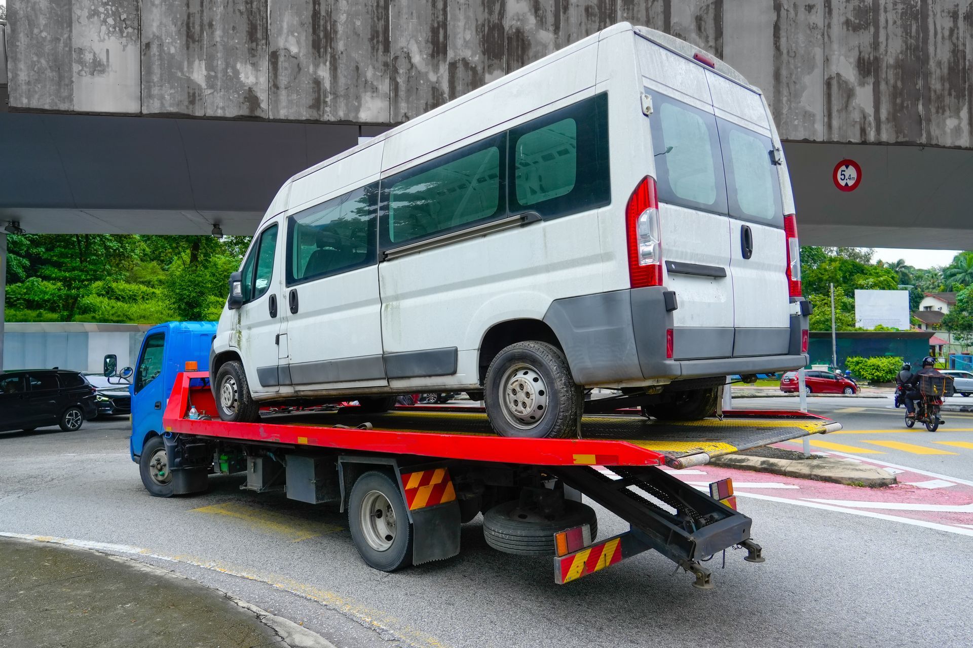 A flatbed tow truck transporting a white van under an overpass on a city road during daytime.