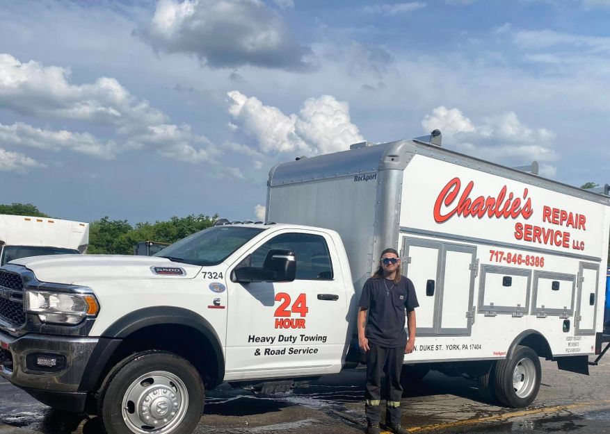 A person stands beside a white repair truck with Charlie's Repair Service on it. Sky is overcast. A person stands beside a white repair truck with Charlie's Repair Service on it. Sky is overcast.
