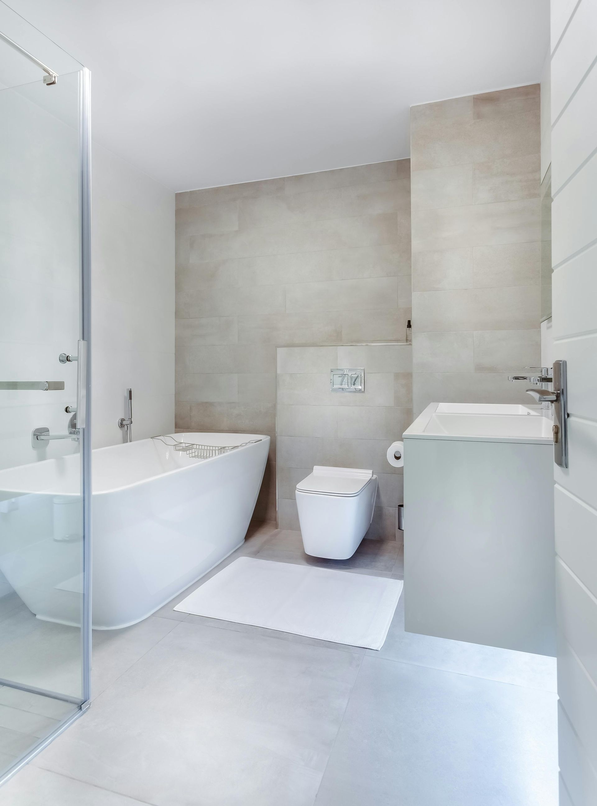 Modern bathroom with white tub, toilet, and sink; beige tiled walls; glass shower door.