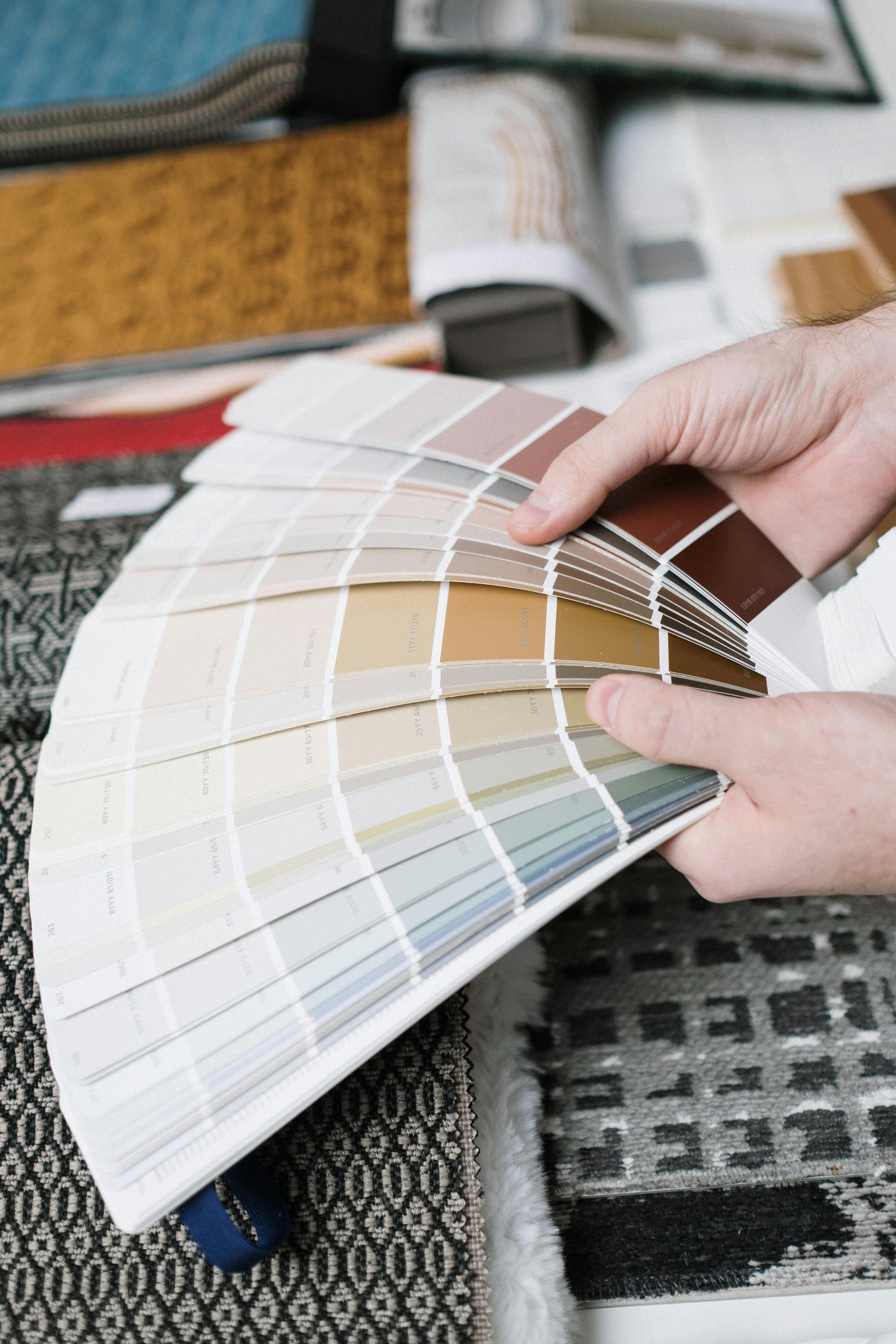 Person holding a color swatch fan, selecting a paint color with fabric samples in the background.