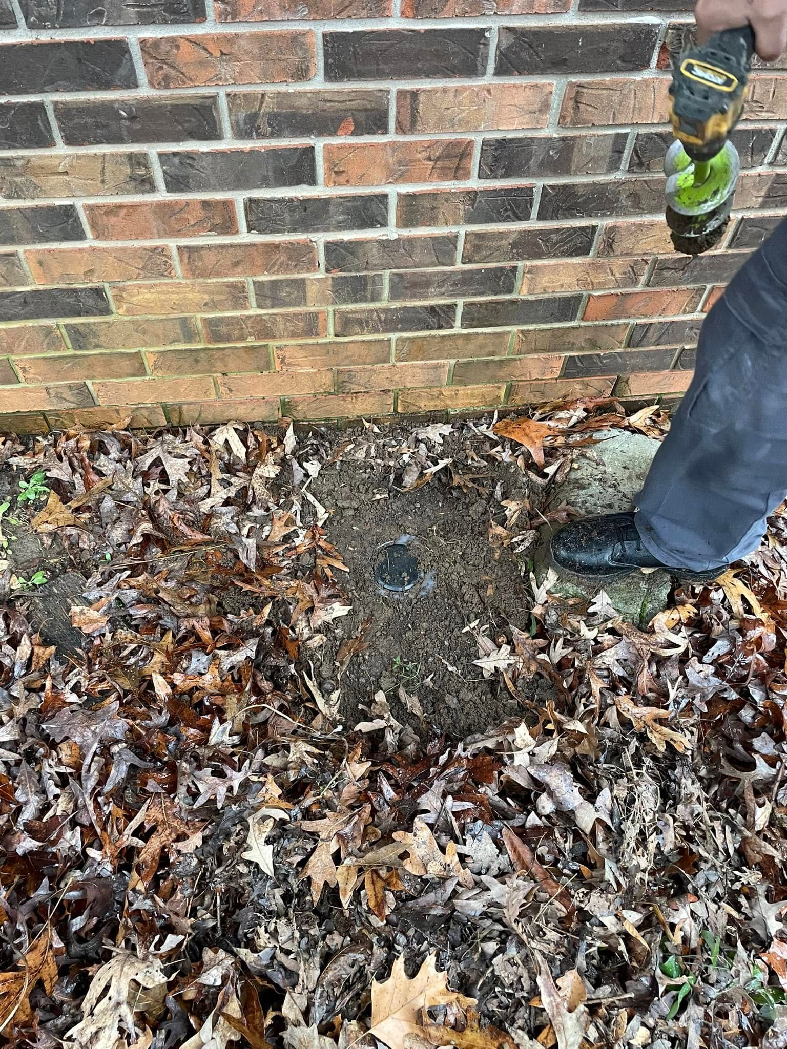A person is using a drill to dig a hole in the ground next to a brick wall.
