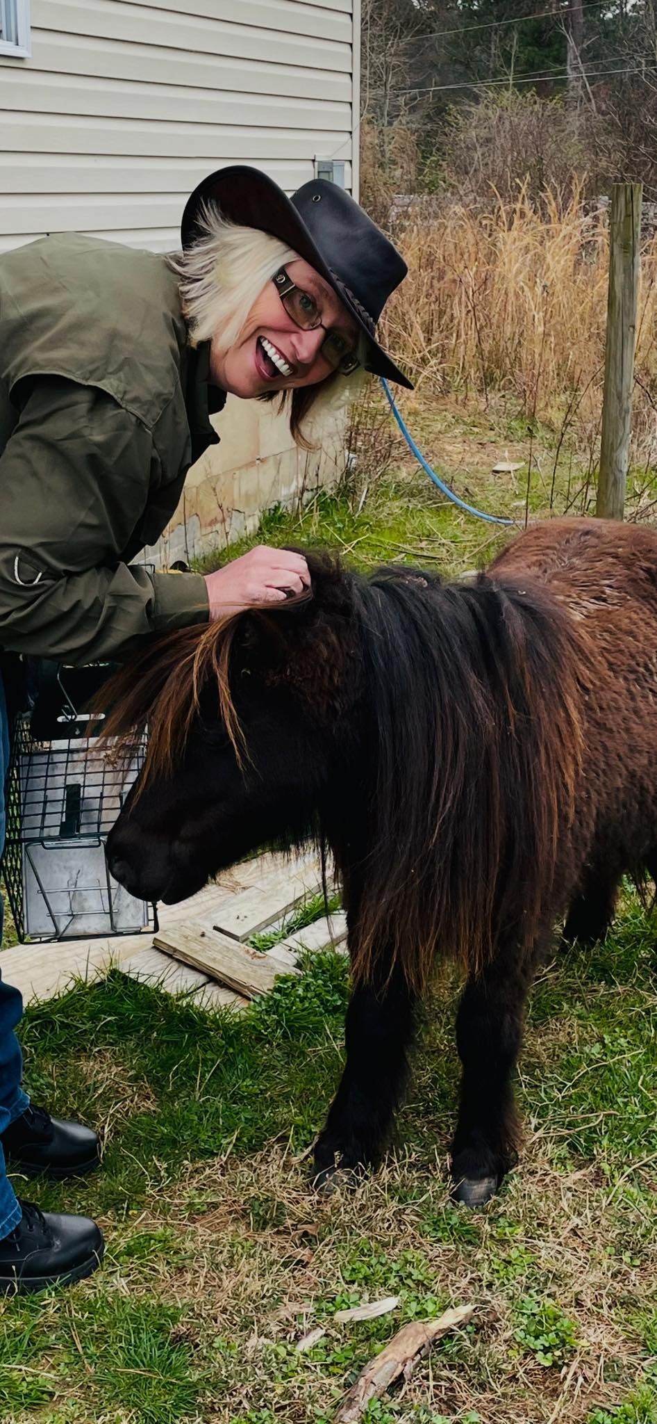 A man in a cowboy hat is petting a small pony in the grass.